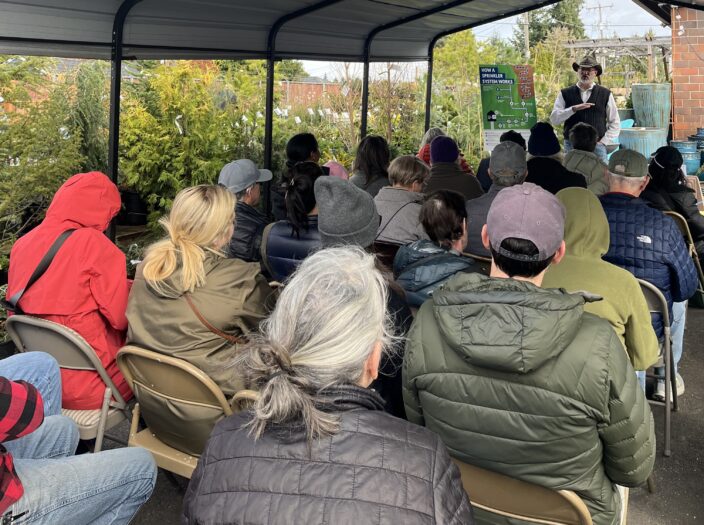 Mark Guthrie teaches a group of people in chairs surrounded by plants at a nursery. 