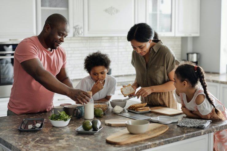 African American family stands around a kitchen island eating snacks. 
